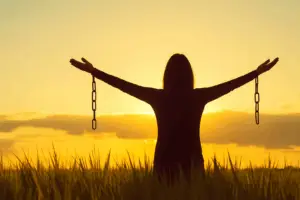 Mujer de espaldas con cadenas rotas en los brazos al amanecer, símbolo de liberación y crecimiento personal.
