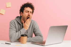 Hombre joven bostezando frente a un portátil con vaso de café y fondo rosa; representa somnolencia diurna por sueño no reparador.