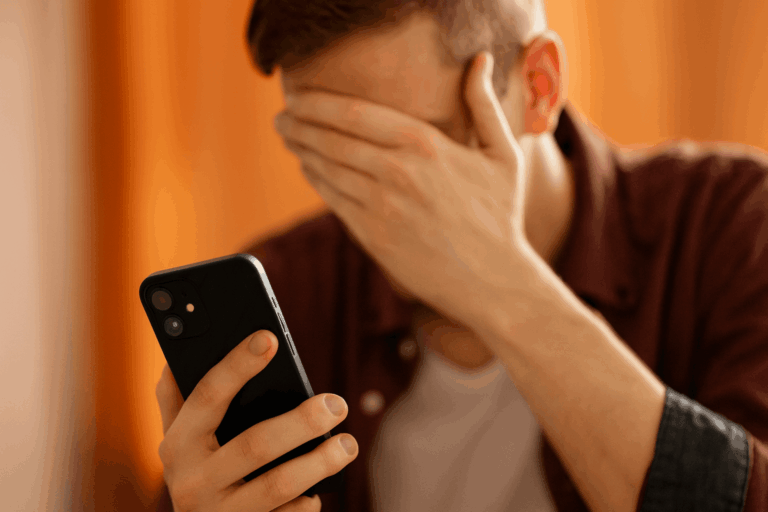 Joven con mano en la cara mirando su smartphone, gesto de estrés, fondo naranja desenfocado.
